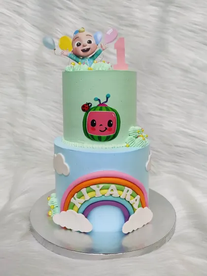 This two-tier Cocomelon cake combines a rainbow theme on the bottom with a JJ topper on top, making it a vibrant centerpiece for a first birthday party.