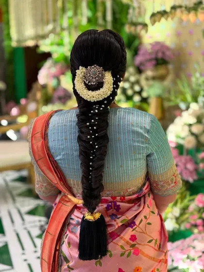 A stunning bridal look captured against a floral backdrop. The fishtail braid is accessorized with a statement silver piece and pearls, creating a beautiful contrast with the bride's colorful saree.