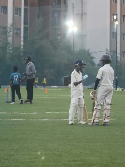 Two young batters confer mid-pitch during a break in their Under-10 practice match. This shows the development of teamwork and strategy from a very young age.