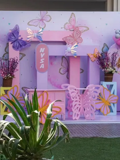 A beautiful butterfly-themed party setup in pastel pink and purple. The decor features a photo-frame style backdrop and whimsical butterfly cutouts.