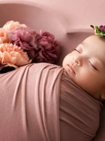A sleeping newborn wrapped in a dusty pink swaddle, with a floral headband and a bouquet of matching flowers.