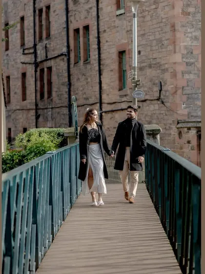 The couple walks across a bridge in Edinburgh, hand-in-hand. This full-length shot captures their stylish coordination and the beautiful stone architecture of the city.