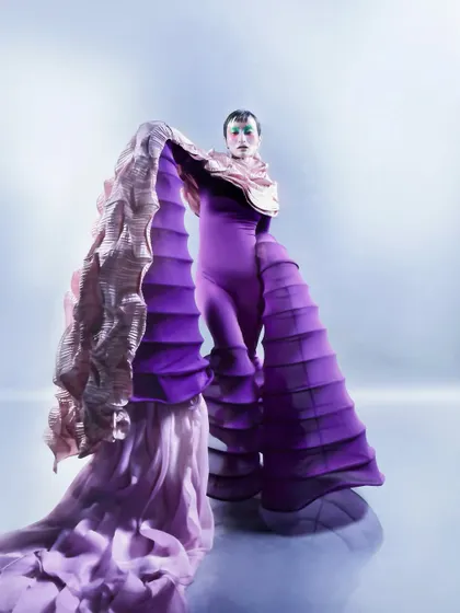 A full-body shot of the model in a sculptural purple and pink ensemble, showcasing the garment's dramatic, accordion-like structure. The pose and lighting emphasize the avant-garde silhouette against a clean, misty background.