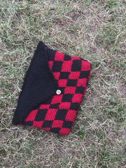 A top-down view of the checkered book sleeve on the grass. It's made from soft yarn to cushion your book.