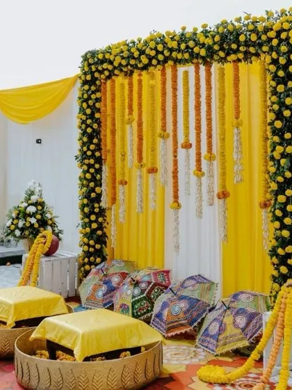 A simple and bright haldi setup with a frame of marigolds against a yellow and white draped background. The traditional brass urlis for seating are a classic touch, and the colorful cushions add a pop of fun.