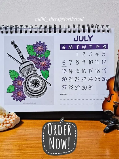 The July page of the calendar, showcasing a detailed violin illustration nestled among purple dahlias. The calendar is spiral-bound and sits perfectly on any desk.