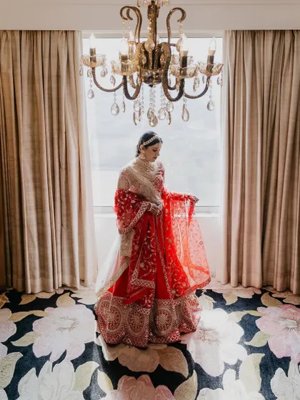 A classic bridal portrait in a beautifully lit room. The chandelier and elegant decor complement the bride's stunning red lehenga, creating a picture of pure grace.