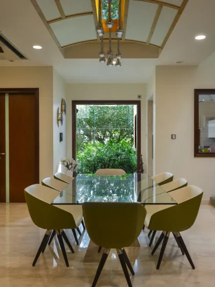 The dining room at Gupta Residence features a large window framing lush greenery, infusing the space with freshness. The ceiling with wooden accents and a modern pendant light adds a touch of elegance.