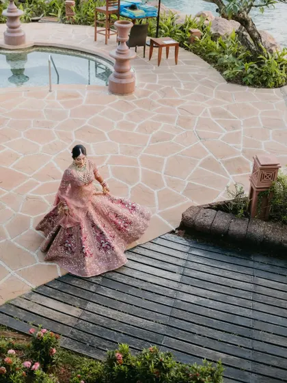 An aerial view of a bride in a pink lehenga twirling by a resort pool, showcasing the beautiful flare of her outfit.