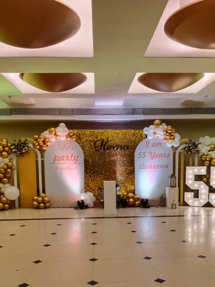This image captures the complete 55th birthday stage setup. The lighting highlights the different textures of the gold shimmer wall, the smooth white panels, and the metallic shine of the balloons.