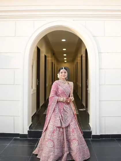 A beautiful full-length shot of the bride in her pastel pink lehenga, framed by a white archway, exuding grace and elegance.