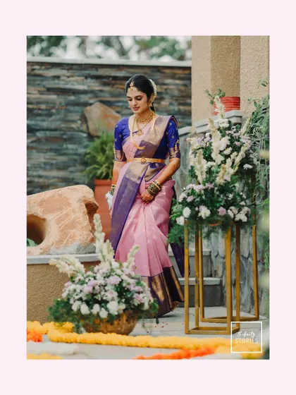 A full-length portrait of the bride descending a staircase, surrounded by floral arrangements. She looks graceful and elegant in her pink and blue silk saree.