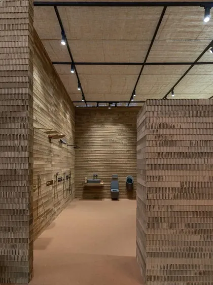 An interior view of the Aquant stall, showcasing bathroom fixtures against walls made of layered cardboard. The minimal, monochrome backdrop ensures the products are the main focus.