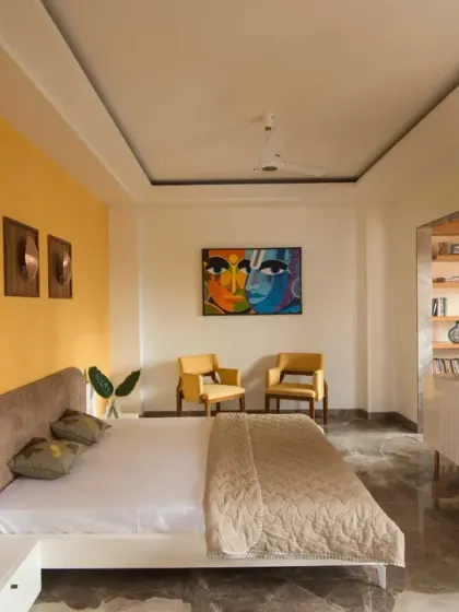 The master bedroom in Kavyam Residence blends warmth and modern elegance. A vibrant yellow accent wall, marble flooring, and a cozy seating corner with bold artwork create a refined, contemporary look.