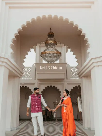 The couple holding hands under a grand chandelier, framed by the arches of their Udaipur wedding venue. A portrait of elegance and love.