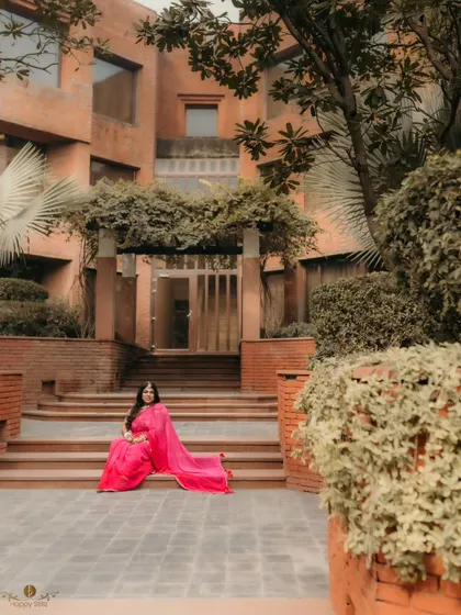 A stunning wide-angle shot of the bride-to-be in a vibrant pink saree, posing on the steps of a beautiful brick building before her haldi ceremony.