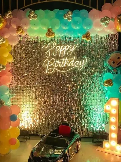 A first birthday setup with a silver shimmer wall and a pastel rainbow balloon arch. The decor is completed with a toy car for the birthday boy and a light-up number '1'.