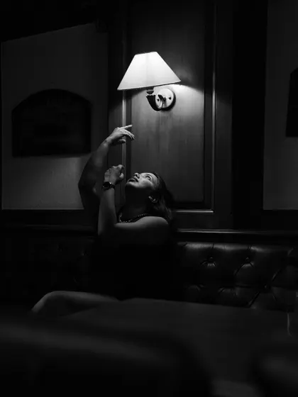 A dramatic black and white portrait of a woman in a dark room, reaching up towards a single wall lamp. The play of light and shadow creates a highly theatrical and artistic composition.