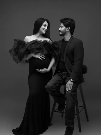 This black and white portrait captures a quiet moment of connection. The expecting mother in a dramatic black ruffled gown looks at her partner with a gentle smile, creating a sophisticated and timeless fine art maternity photograph.