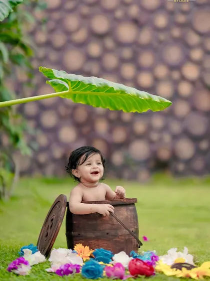 A happy baby enjoying the outdoors, peeking out of a wooden barrel under the shade of a large leaf.