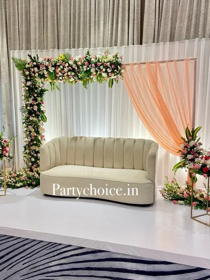A simple yet elegant reception stage setup. The decor features a modern sofa on a white platform, framed by an L-shaped floral arch and accented with a single peach-colored drape for a touch of softness.