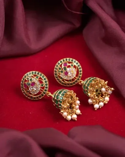 These unique earrings feature a peacock motif stud with green and ruby-colored stones, leading to a small, green-enameled jhumka drop with pearls.