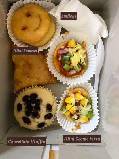 A snack box designed to bring smiles, especially for kids' parties. It includes a Choco-Chip Muffin, Mini Veggie Pizza, a Mini Samosa, and fun potato Smileys.