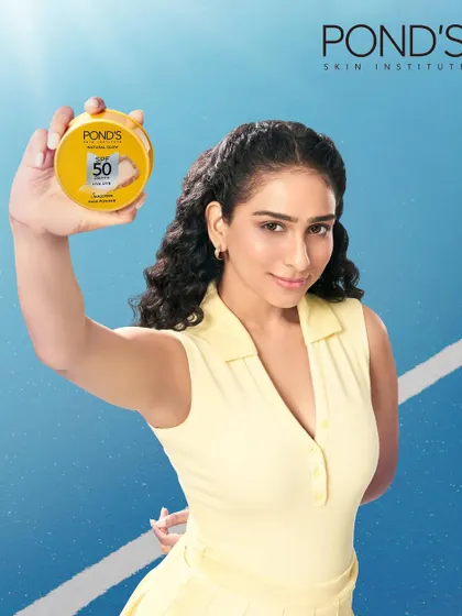 An active lifestyle photograph for the Pond's face powder campaign. The model is dressed in athletic wear against a tennis court background, showcasing the product's suitability for an on-the-go, active life.