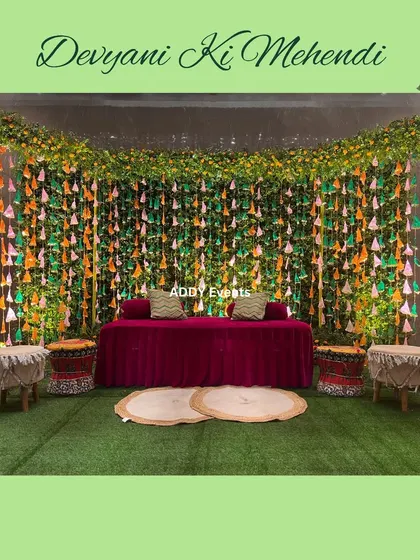 A vibrant Mehndi stage with a backdrop of green foliage and colorful hanging tassels, creating a festive and playful setting.
