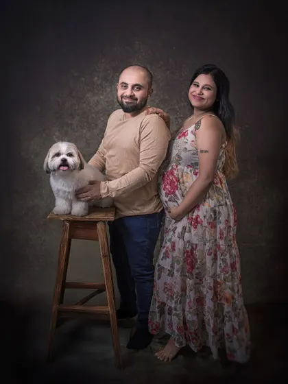 An expecting couple poses with their beloved Shih Tzu in a classic studio portrait. The dog sits proudly on a stool, an important member of the growing family.