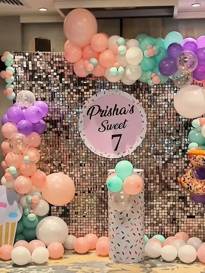 A "Sweet 7" birthday party with a candy and donut theme. This setup features a silver shimmer wall, pastel balloons, and giant cupcake and donut props.