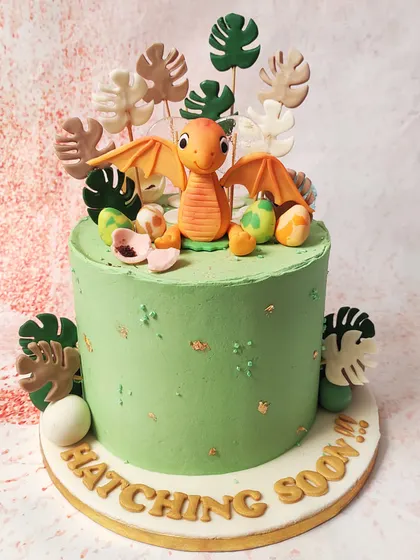 We announce a special arrival with this "Hatching Soon" dinosaur cake. A cute baby pterodactyl sits among its eggs and tropical leaves on a beautiful green cake, making it a unique and adorable theme for a baby shower.
