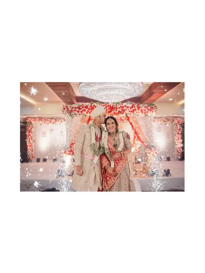 A grand, celebratory moment. The groom kisses his bride's cheek as cold pyrotechnics go off around them, creating a spectacular and festive wedding stage shot.