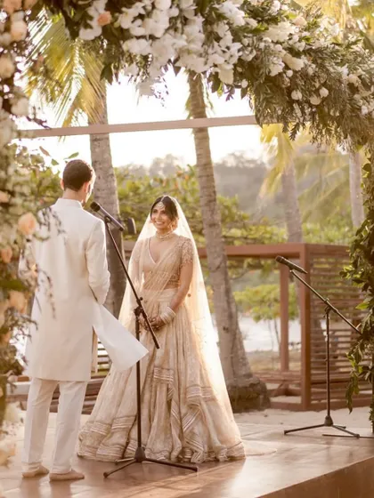 Exchanging vows under a beautiful floral arch. The couple's matching ivory outfits create a serene and romantic atmosphere for their beach wedding.