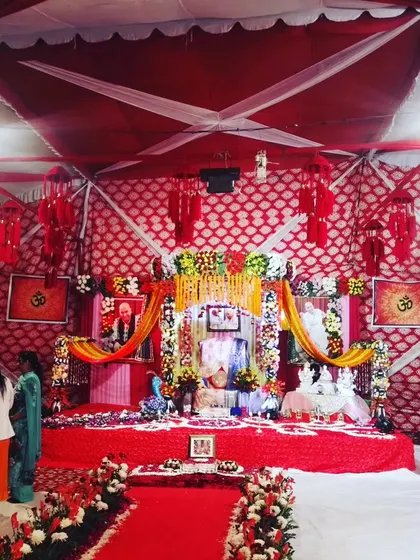 A full view of a large-scale satsang decoration inside a tent. The main stage is elaborately decorated with flowers, lighting, and portraits, creating a divine focal point for the event.