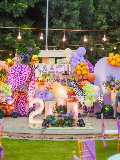 A vibrant and bold jungle theme for a second birthday, featuring a large stage with realistic animal props, including a lion and a purple-spotted giraffe, set in an outdoor space.