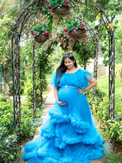 A beautiful solo portrait of a mom-to-be in a blue ruffled gown and tiara, standing under a floral arch in a garden.