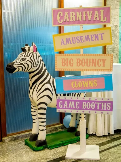 A pastel-colored sign for a carnival party, featuring a zebra prop and directions to 'Amusement', 'Clowns', and 'Game Booths'.