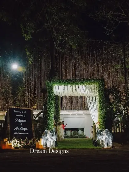 The grand entrance to a nighttime wedding. A leafy archway, a personalized welcome sign, and two decorative elephants create a memorable and traditional welcome for guests.