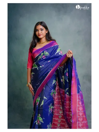 A studio shot for a saree brand. The model's pose displays the full length and vibrant colors of the blue and pink silk saree against a textured studio background.