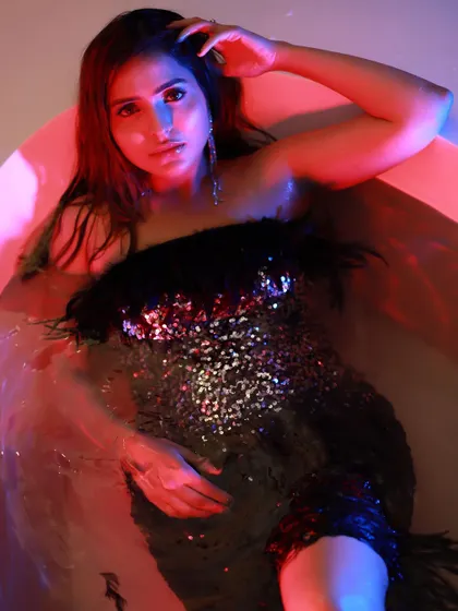 An artistic shot of a model in a feathered sequin dress, sitting in a bathtub with colored lighting. This demonstrates my creative and conceptual capabilities.
