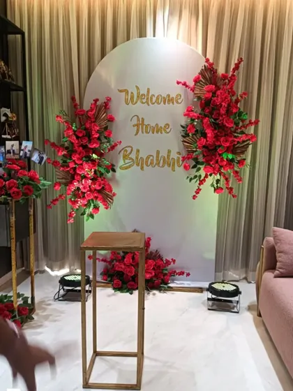 A sweet "Welcome Home Bhabhi" decoration. The white arched backdrop with gold lettering is flanked by bold red floral arrangements.