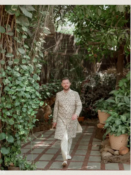 The groom walks confidently down a green pathway, his floral sherwani standing out.