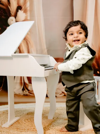 A happy little gentleman, dressed in a vest, stands and plays at the miniature piano in a warm, indoor setting.