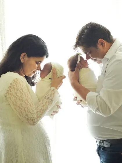 A special welcome for newborn twins. Both parents hold one of their precious babies, gazing at each other in this light-filled, emotional family portrait.