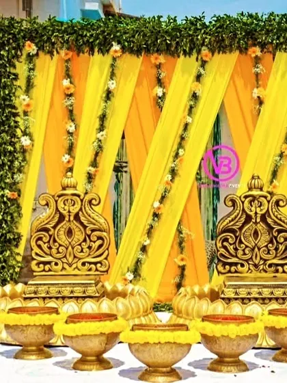 A beautiful Haldi setup with ornate golden chairs, a backdrop of yellow drapes and green foliage, and traditional brass bowls.