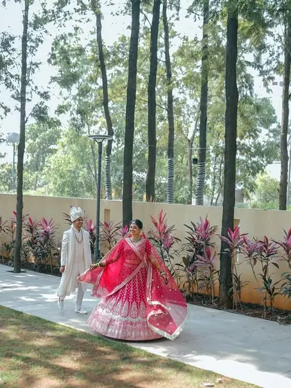 A walk to remember. The bride's vibrant pink lehenga flows beautifully as she and her groom stroll through a path lined with tall, majestic trees, creating a cinematic and artistic wedding portrait.