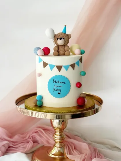 Another sweet "Welcome Home Baby" cake. This design features a teddy bear topper, a blue name plaque, and a colorful pom-pom garland.
