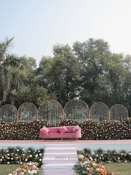 A full view of a beautiful outdoor stage setup. The backdrop features golden arches with hanging floral strings, a base of colorful flowers, and a chic pink velvet sofa, creating a fresh and modern look for a day event.