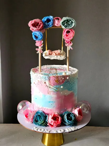 A beautiful baby shower cake with a working swing. The watercolor-effect buttercream in pink and blue is complemented by paper flowers and a tiny, sleeping baby in a swinging cradle.